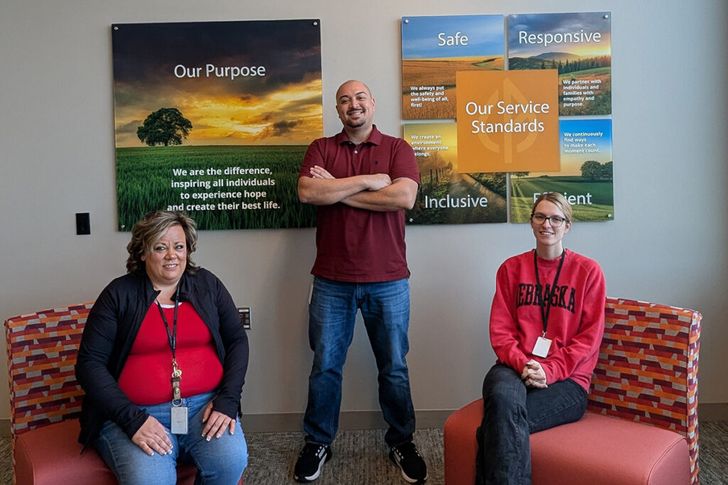 Three Community Alliance staff members posing in an office lobby beneath ‘Our Purpose’ and ‘Our Service Standards’ wall displays.