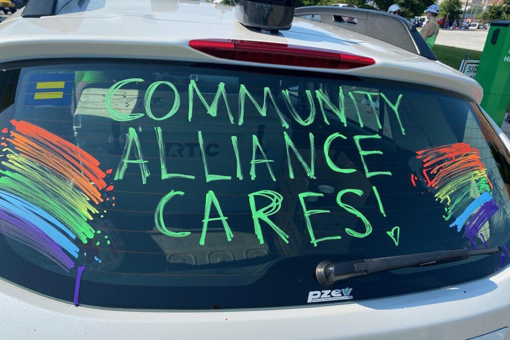 Car rear window painted with the words ‘Community Alliance Cares!’ surrounded by rainbow designs.
