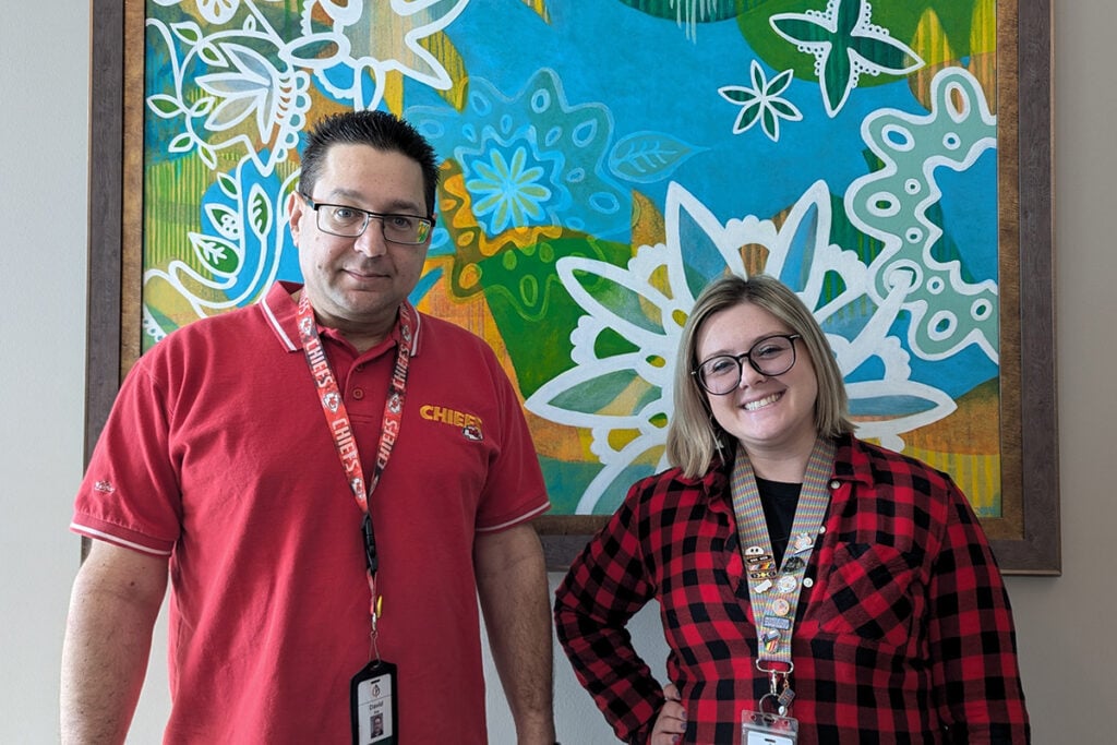 Two Community Alliance staff members standing in front of a colorful mural, smiling at the camera.