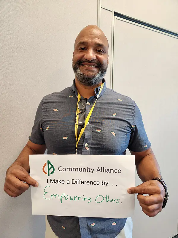 Smiling man holding a sign that reads ‘Community Alliance – I Make a Difference by… Empowering Others.”