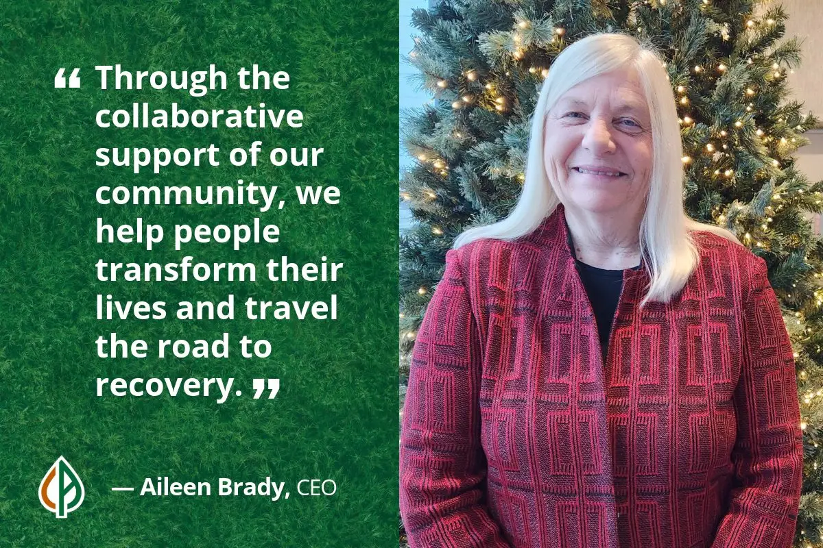 Photo of Aileen Brady, CEO, smiling in front of a lit holiday tree, alongside a quote that reads: “Through the collaborative support of our community, we help people transform their lives and travel the road to recovery.”
