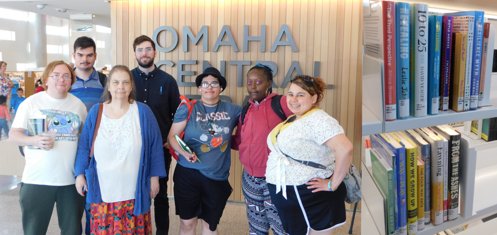 A group of six adults and one staff member pose together inside the Omaha Central Library. They are standing in front of a wood-paneled wall with large “OMAHA CENTRAL” lettering. The group is smiling and dressed casually, some carrying books, notebooks, or backpacks. A library bookshelf filled with books is visible in the foreground on the right, creating a welcoming library setting.