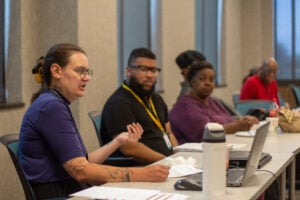 Kendra, who has tattoos and wears glasses, animatedly gestures while speaking to onlooking consumer council members at a conference room table.