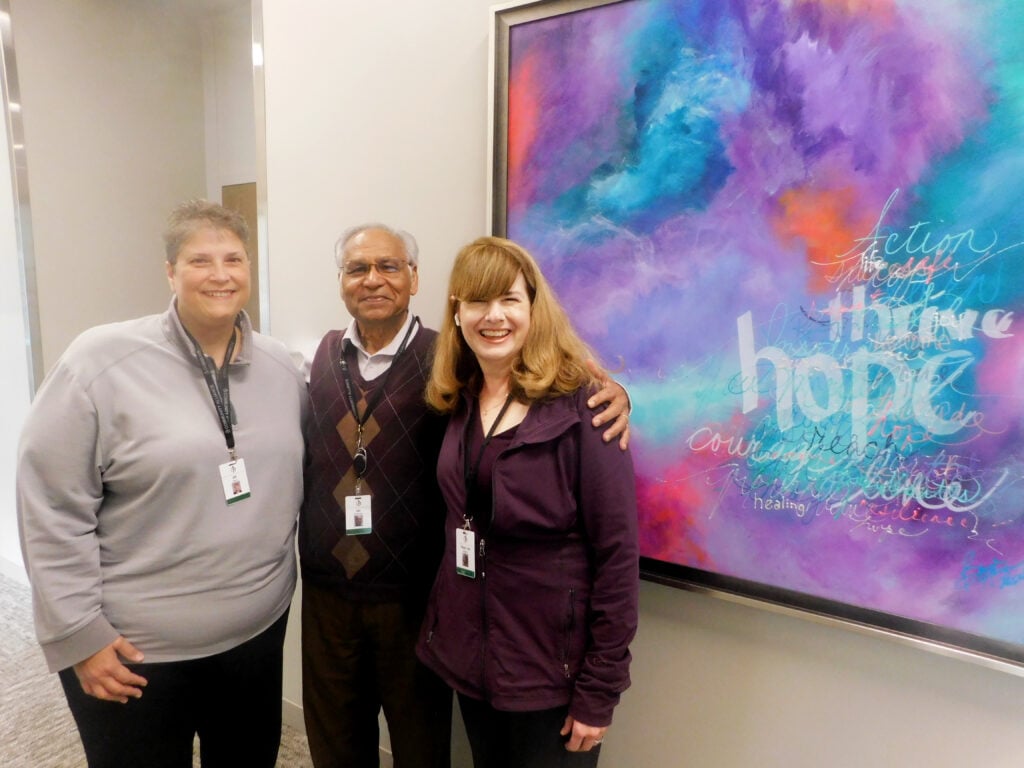 Three members of the Family Support and Education team, Jill Davis, Dr. Jai Sookram and Therese Vaughn, standing and smiling next to each other near a vibrant painting that says thrive, hope and other encouraging words