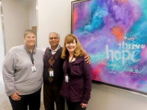 Three members of the Family Support and Education team, Jill Davis, Dr. Jai Sookram and Therese Vaughn, standing and smiling next to each other near a vibrant painting that says thrive, hope and other encouraging words